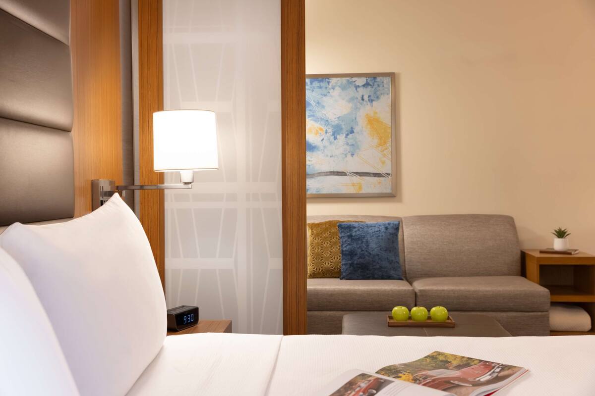 A hotel room with a bed, a nightstand with a clock, a sofa, a wall-mounted lamp, and a painting.