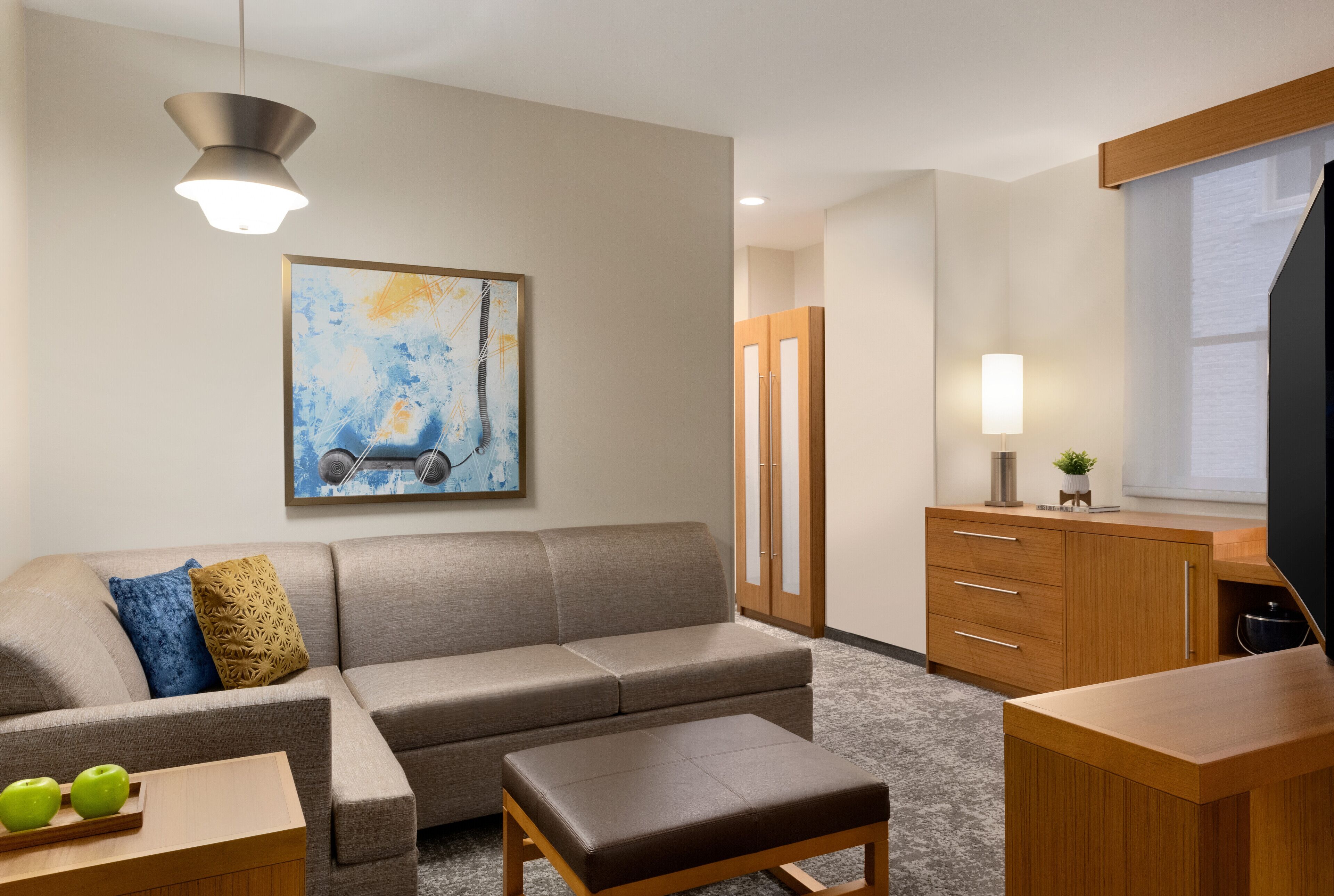 Quarto de hotel moderno com sofá, escrivaninha de madeira e armário. Há uma pintura na parede e um abajur na escrivaninha.