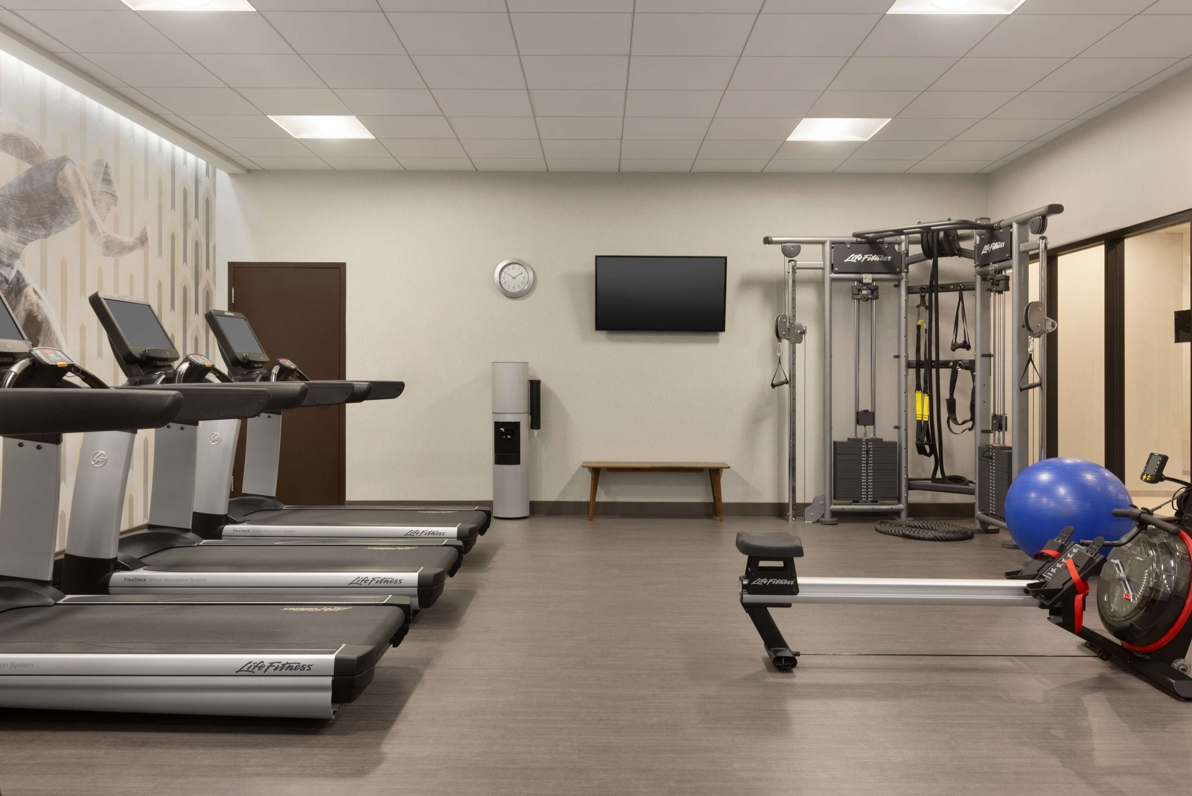 Un gimnasio con cintas de correr, una bicicleta estática, una máquina de remo y un televisor montado en la pared.