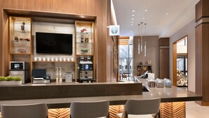A modern bar area with a flat-screen TV, coffee machines, and a variety of snacks. There are three bar stools and a wooden herringbone floor pattern. - Hyatt Place Houston Downtown (Houston)