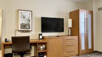 A hotel room with a wooden desk, a chair, a television, a lamp, a wardrobe, and a framed picture on the wall.