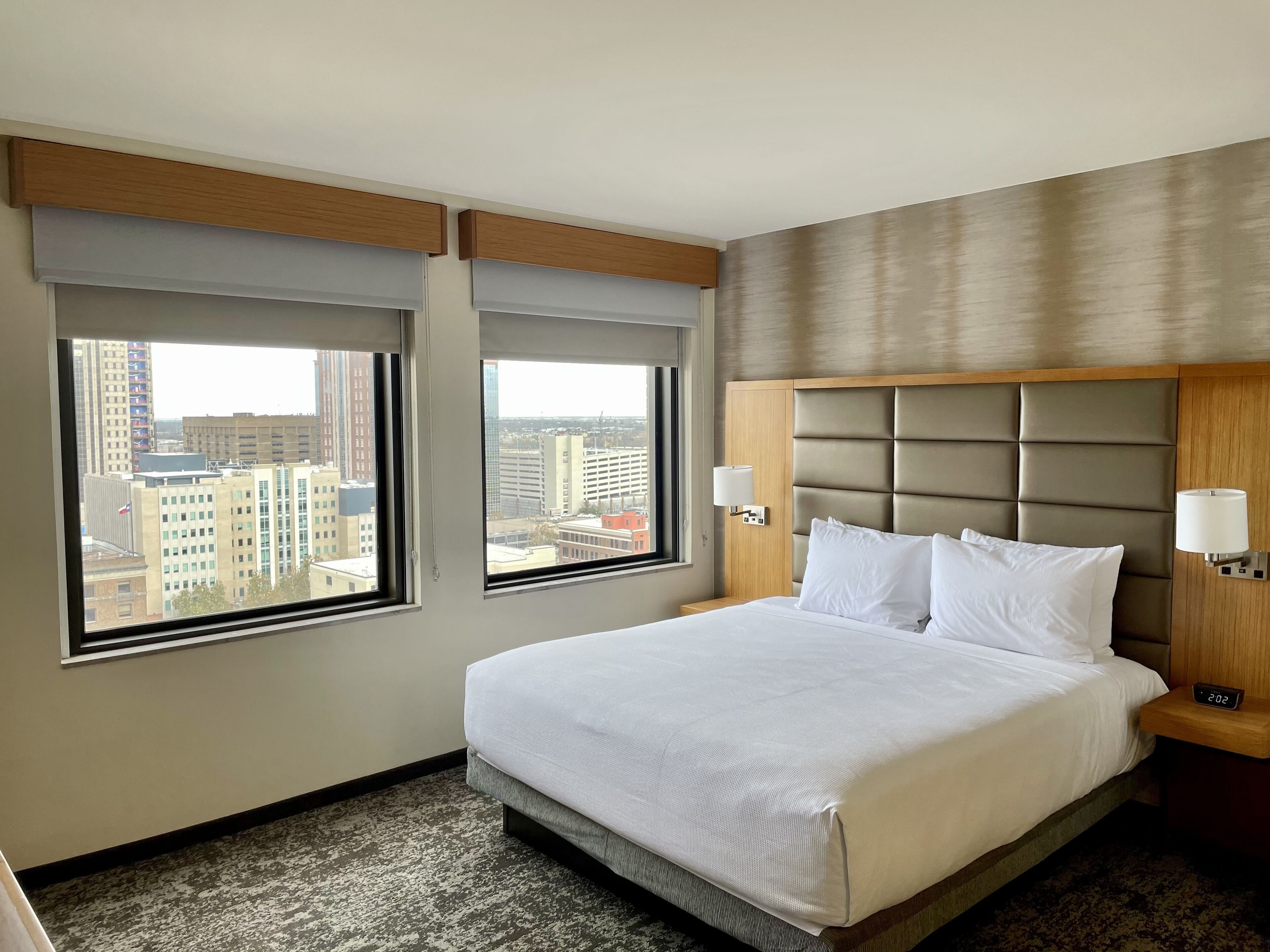A hotel room with a large bed, two windows, a city view, a headboard, and a bedside table with a lamp.