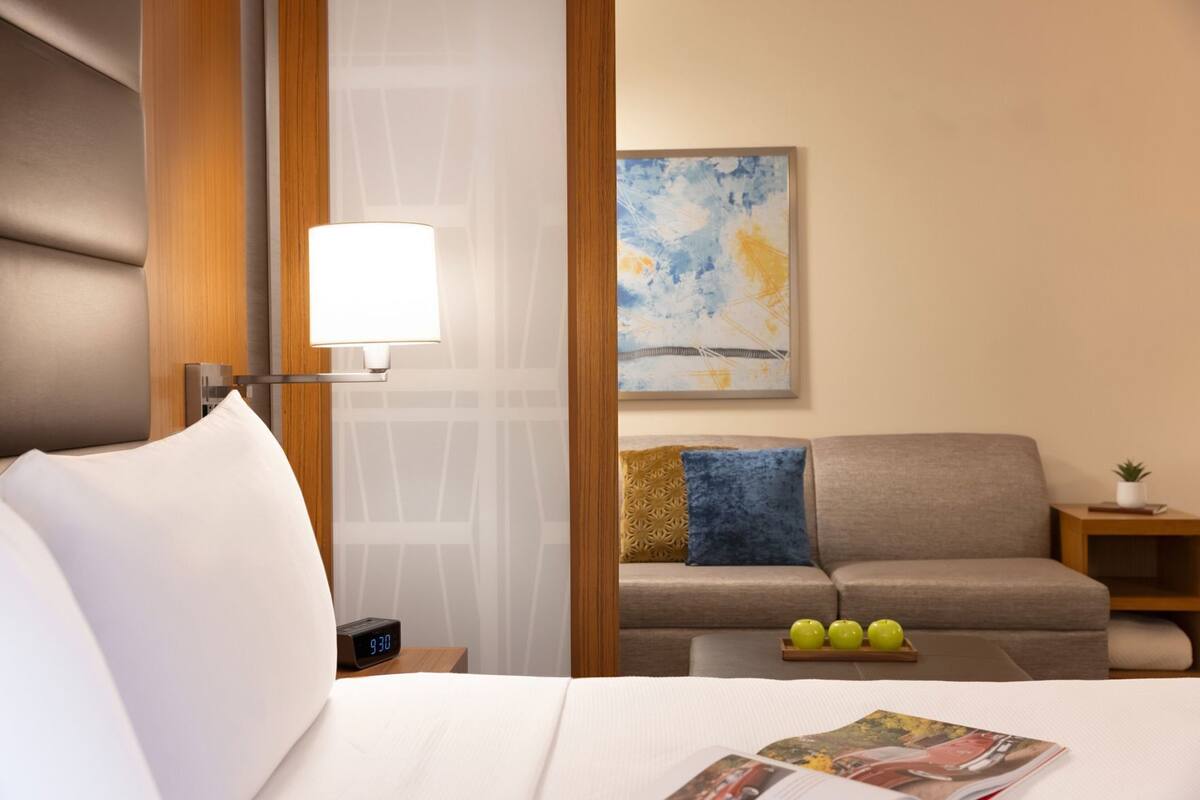 A hotel room with a bed, a bedside table with a clock, a lamp, a sofa with cushions, and a painting on the wall.