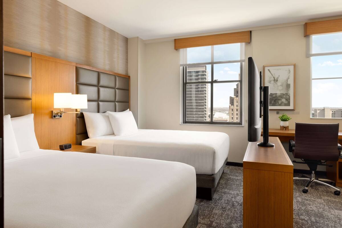 A hotel room with two beds, a desk, a television, and a window with a city view.