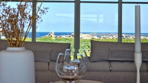 A room with a large window overlooking a rocky coastline, a sofa, a vase with dried flowers, and a wine glass. - Villa Frouden, Beach, sea view, jacuzzi (Plouescat)