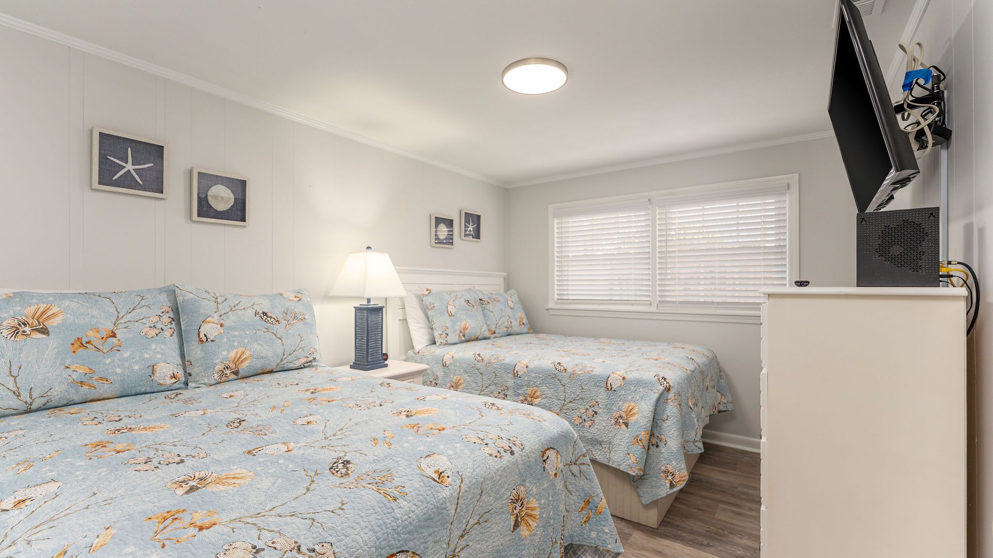 Une chambre avec deux lits, une table de chevet, une lampe, un téléviseur et une fenêtre avec des stores.
