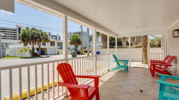 Un espacio al aire libre techado con barandilla blanca, sillas tipo Adirondack rojas y turquesas, y vista a una calle residencial con palmeras y edificios.