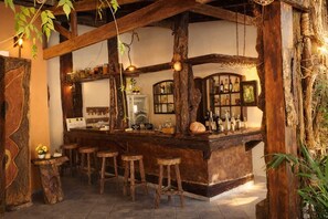 Zona de bar rústica con vigas de madera, mostrador, taburetes y estantes con botellas.