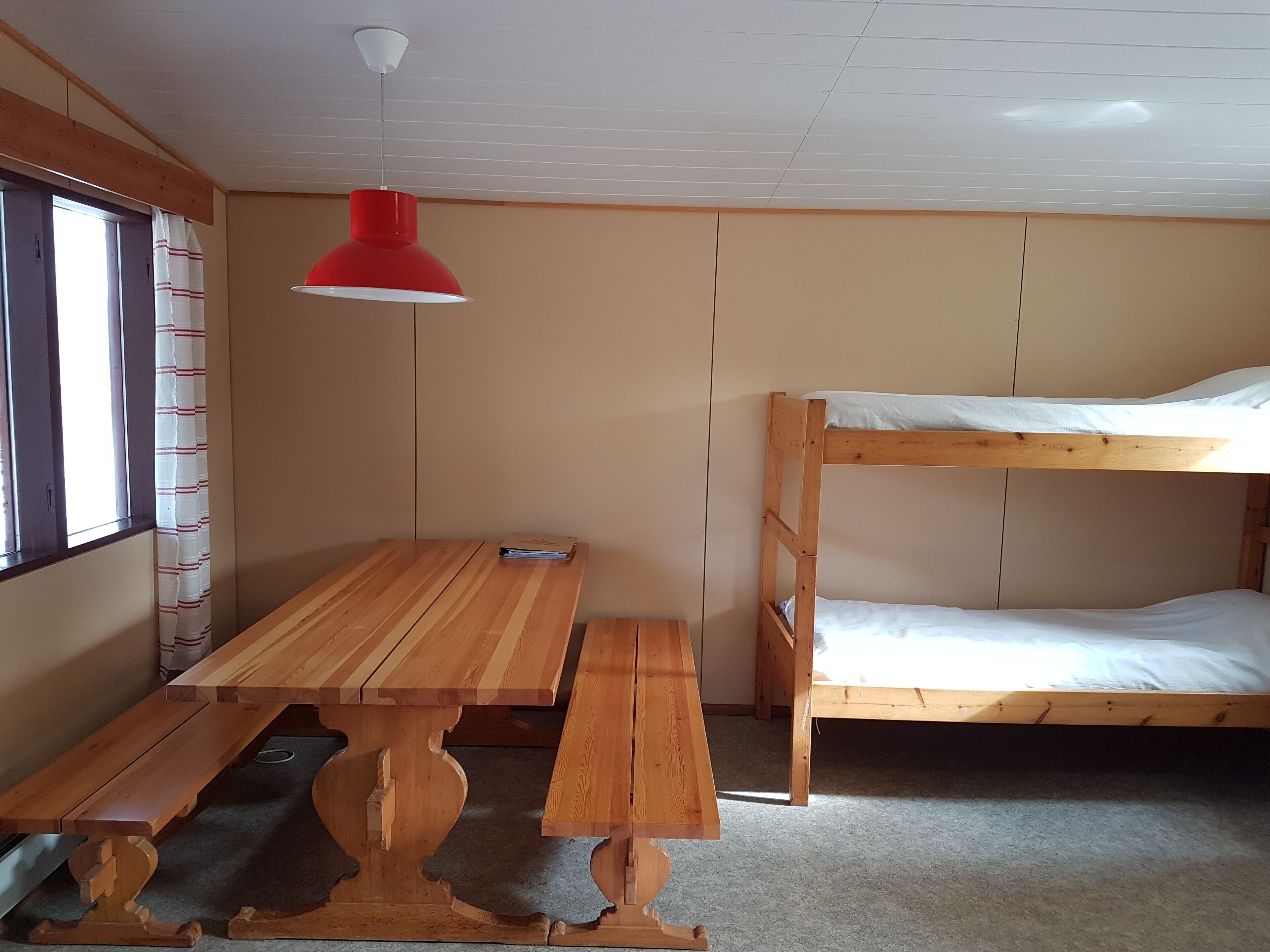 Un lit superposé avec une structure en bois et une literie blanche, une table en bois avec un banc, et une lampe suspendue rouge.