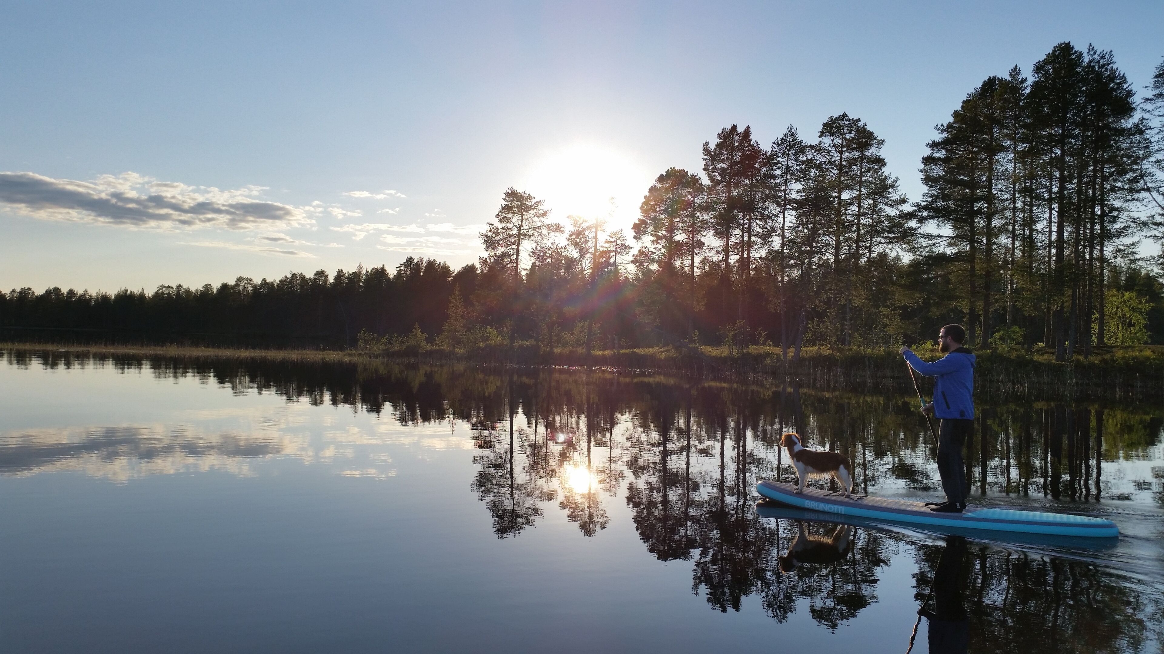 A person on a paddleboard in a calm lake with trees in the background and the sun setting.