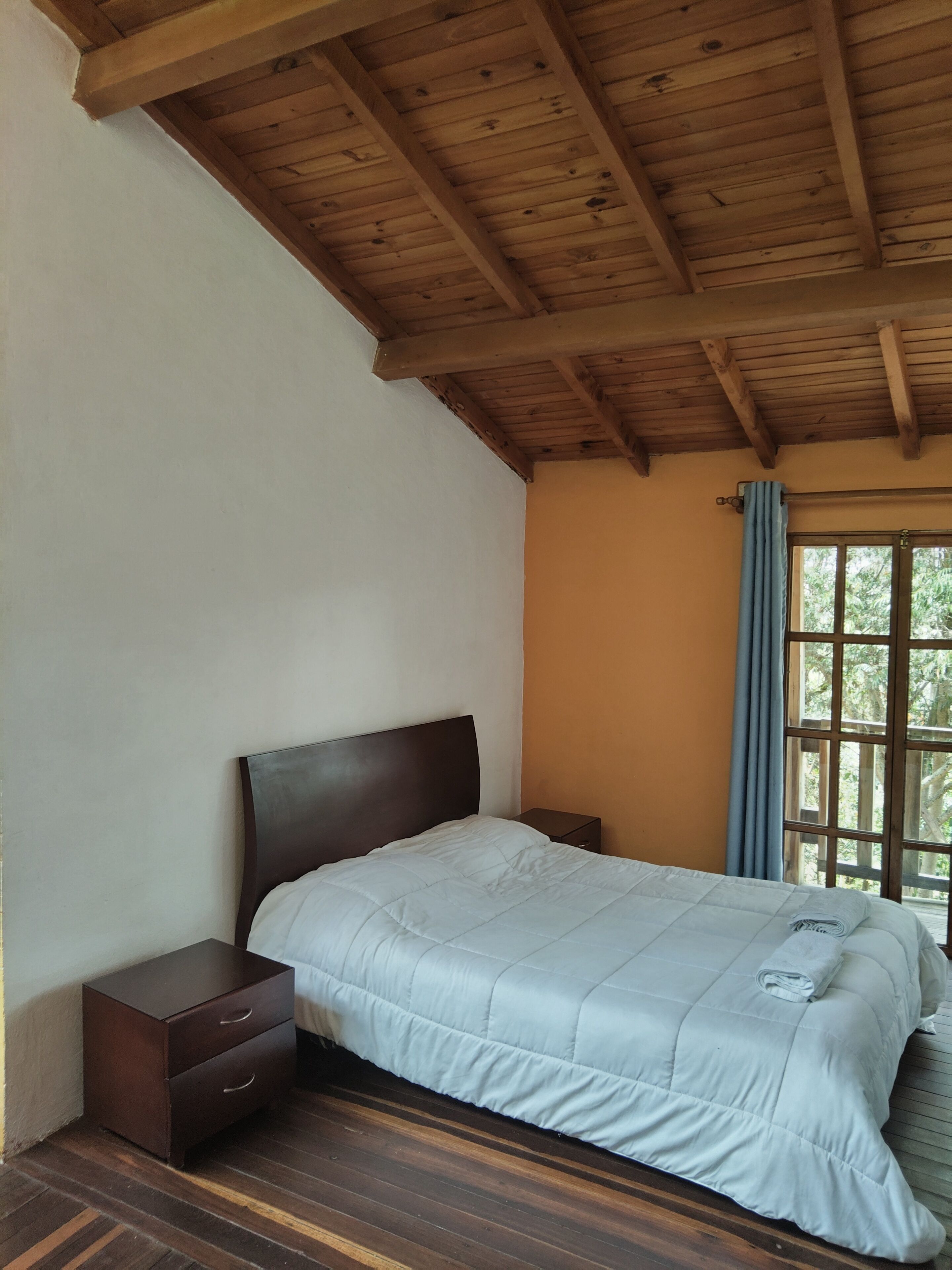 A bedroom with a wooden ceiling, a bed with a white duvet, a nightstand, and a window with a blue curtain.