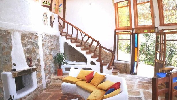 Un interior rústico con chimenea de piedra, una escalera de madera y un sofá blanco con cojines de colores.