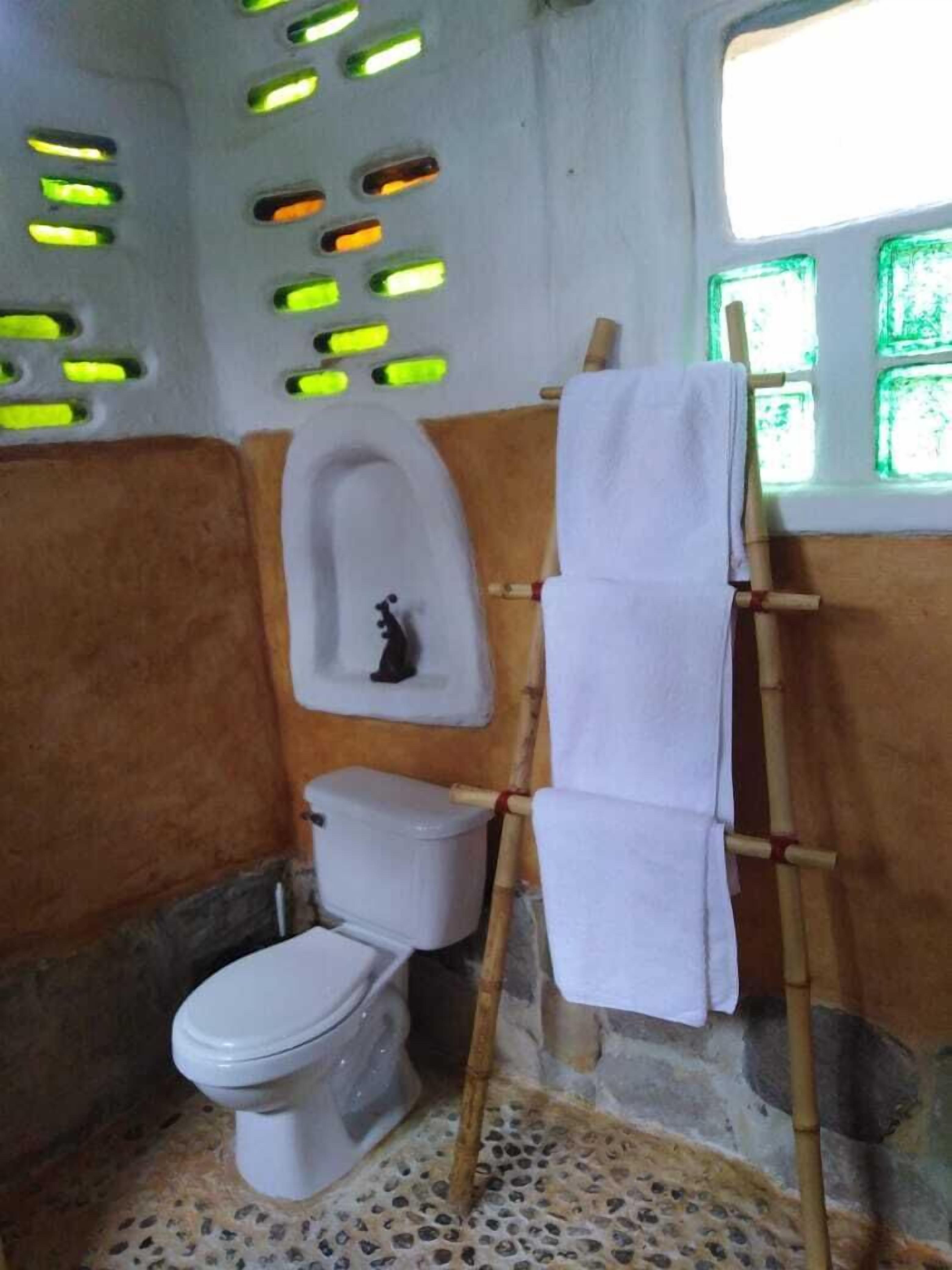Un baño con inodoro, un estante pequeño con una figurina, toallero y una ventana con vidrio tintado de verde.