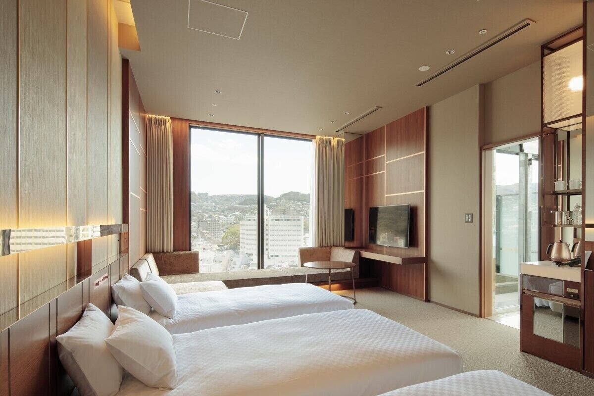 Habitación de hotel con dos camas, televisión, escritorio y vista a la ciudad.