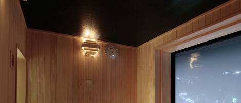 A sauna with wooden walls, a bench, a clock, and a large screen displaying a cityscape.
