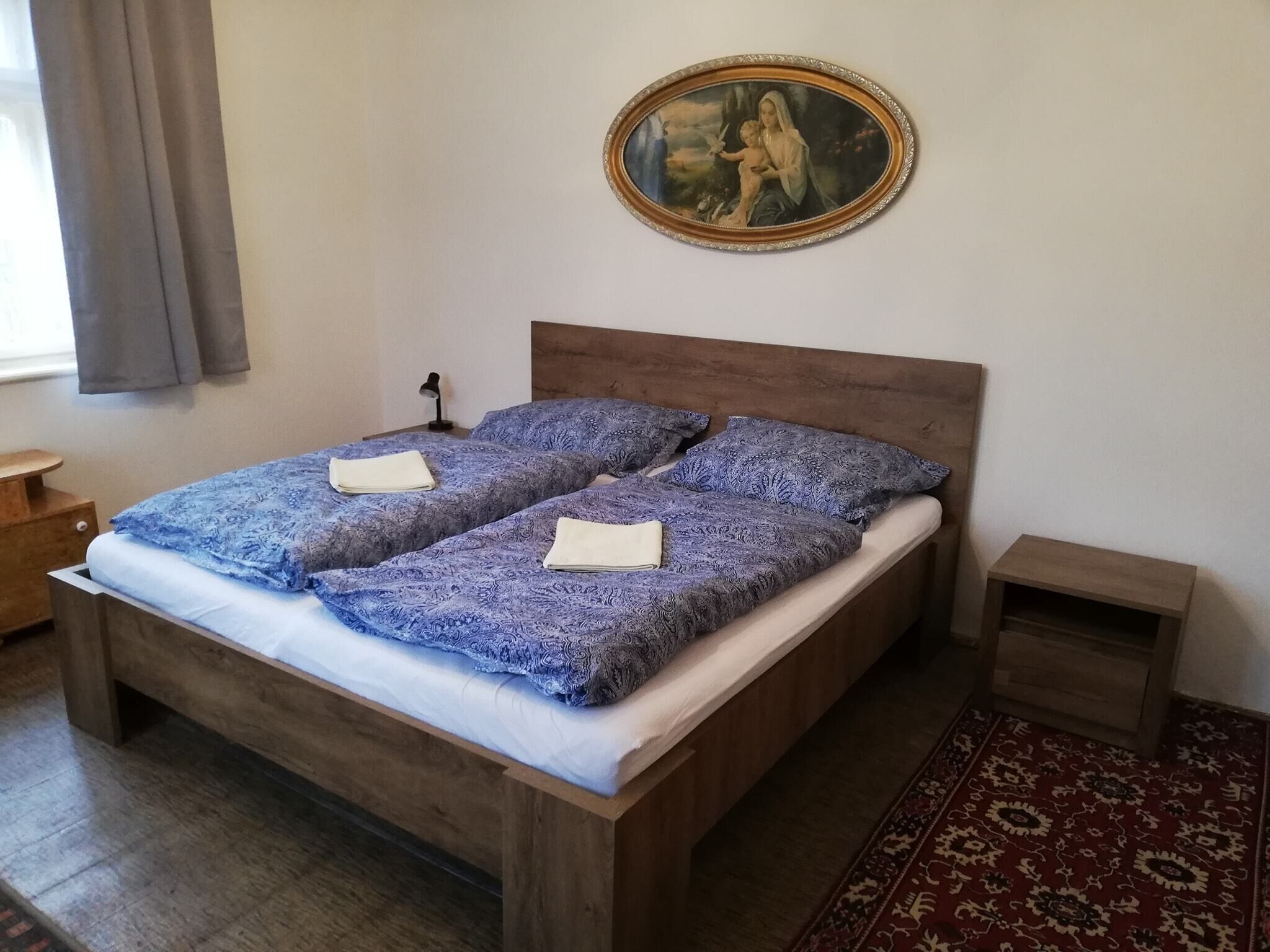 Een tweepersoonsbed met beddengoed met patroon, een houten hoofdbord, een nachtkastje met een lamp en een ingelijst schilderij aan de muur.