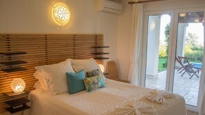 A bedroom with a wooden headboard, white bedding, and decorative pillows. There is a nightstand with a lamp and a small table. The room has a sliding glass door leading to an outdoor area with a chair and a table. - Villa Bethany (Kalkan)