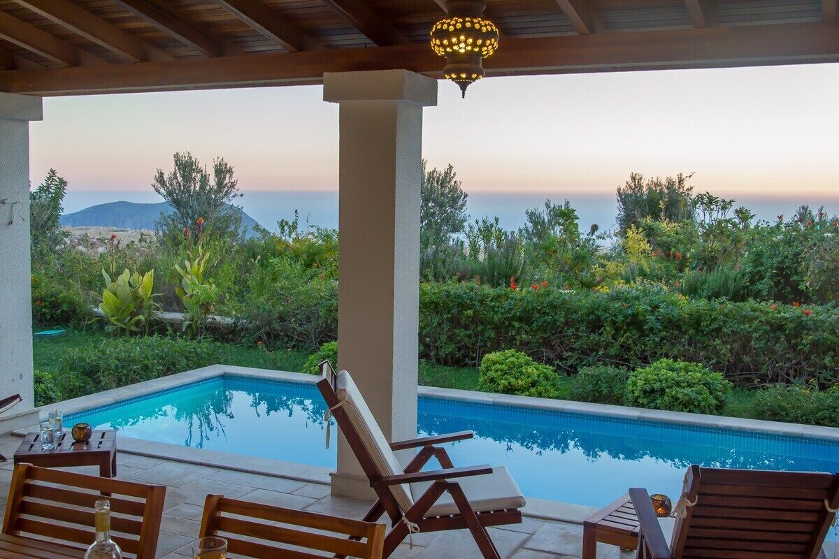 Un espace aménagé au bord de la piscine, avec vue sur la mer, comprenant des chaises longues en bois et une pergola agrémentée d’un luminaire.