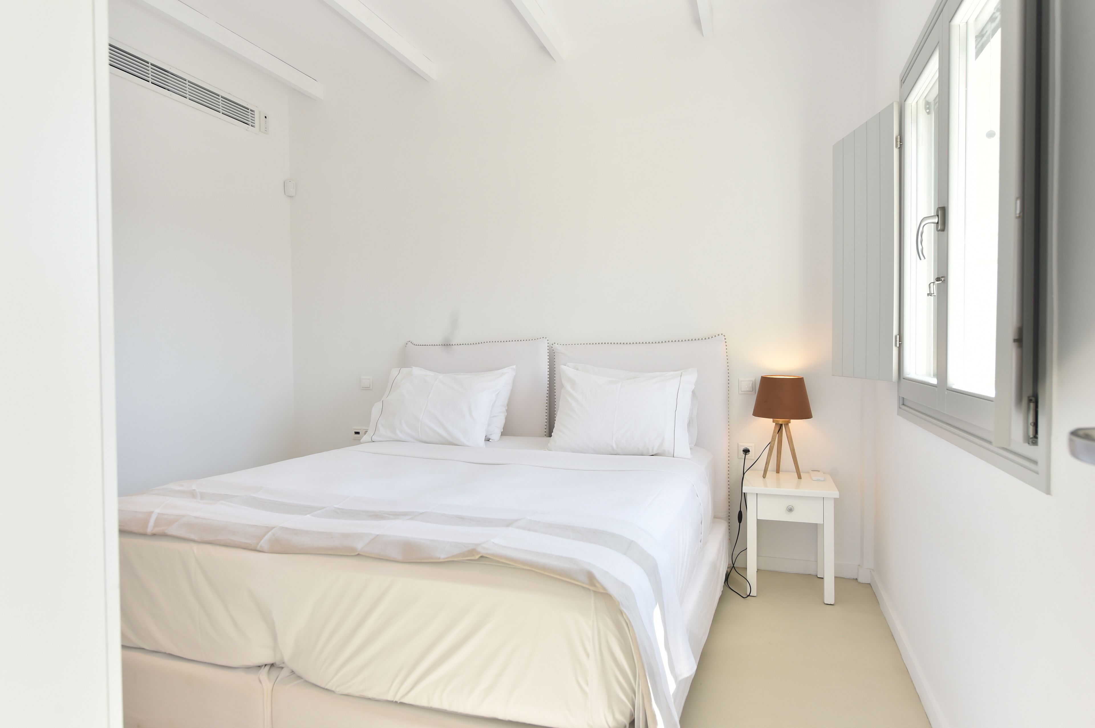 A white bedroom with a bed, bedside table, lamp, and window.