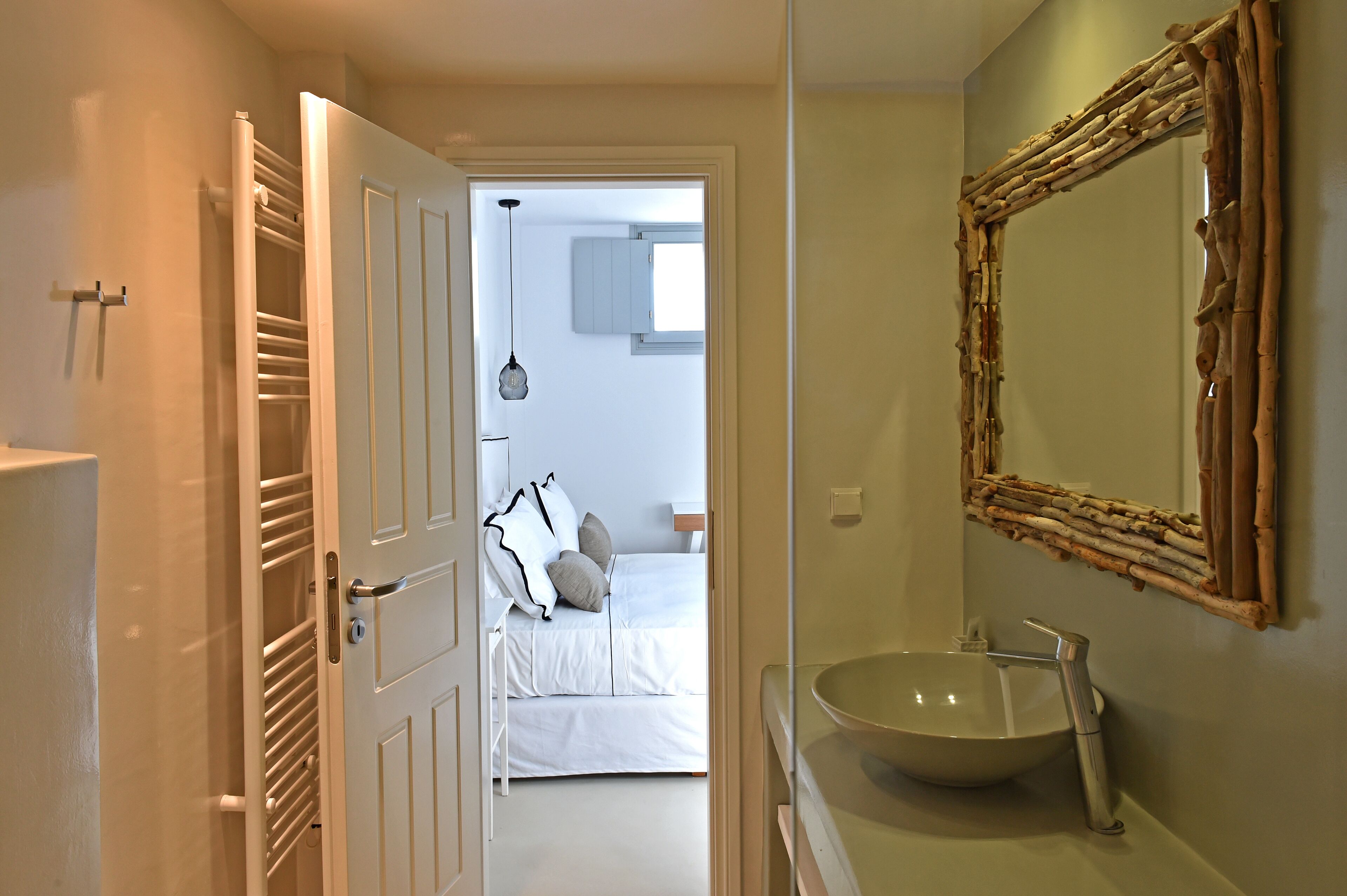 A bathroom with a large mirror, a sink, and a door leading to a bedroom with a bed and a hanging light fixture.