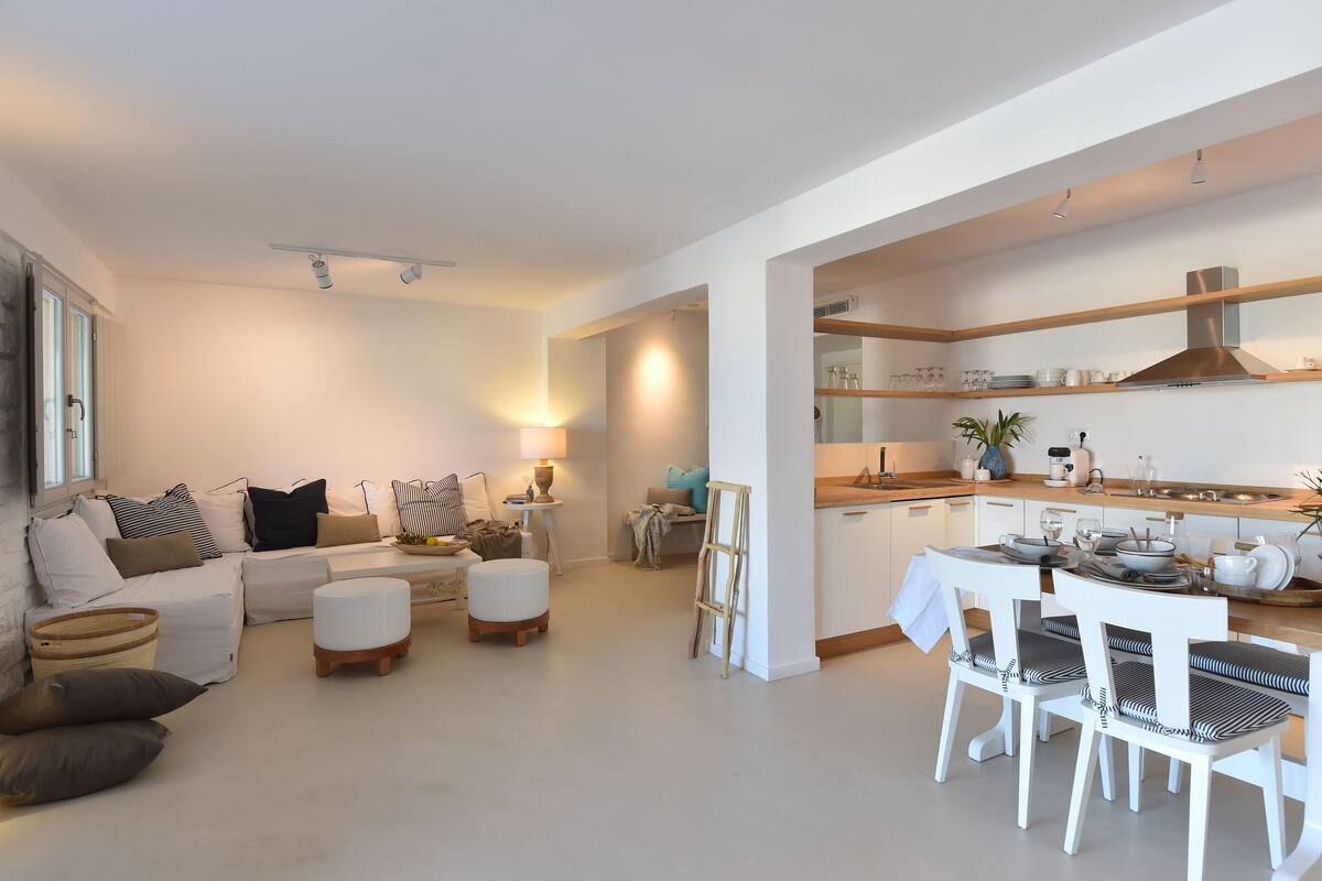 Ein modernes Wohnzimmer mit einer Couch, Hocker und einem Essbereich mit weißen Stühlen und einem Tisch.