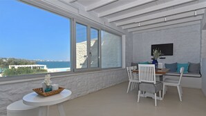 Uma varanda com parede de pedra branca, mesa de jantar com cadeiras, televisão fixada na parede e vista para o mar.