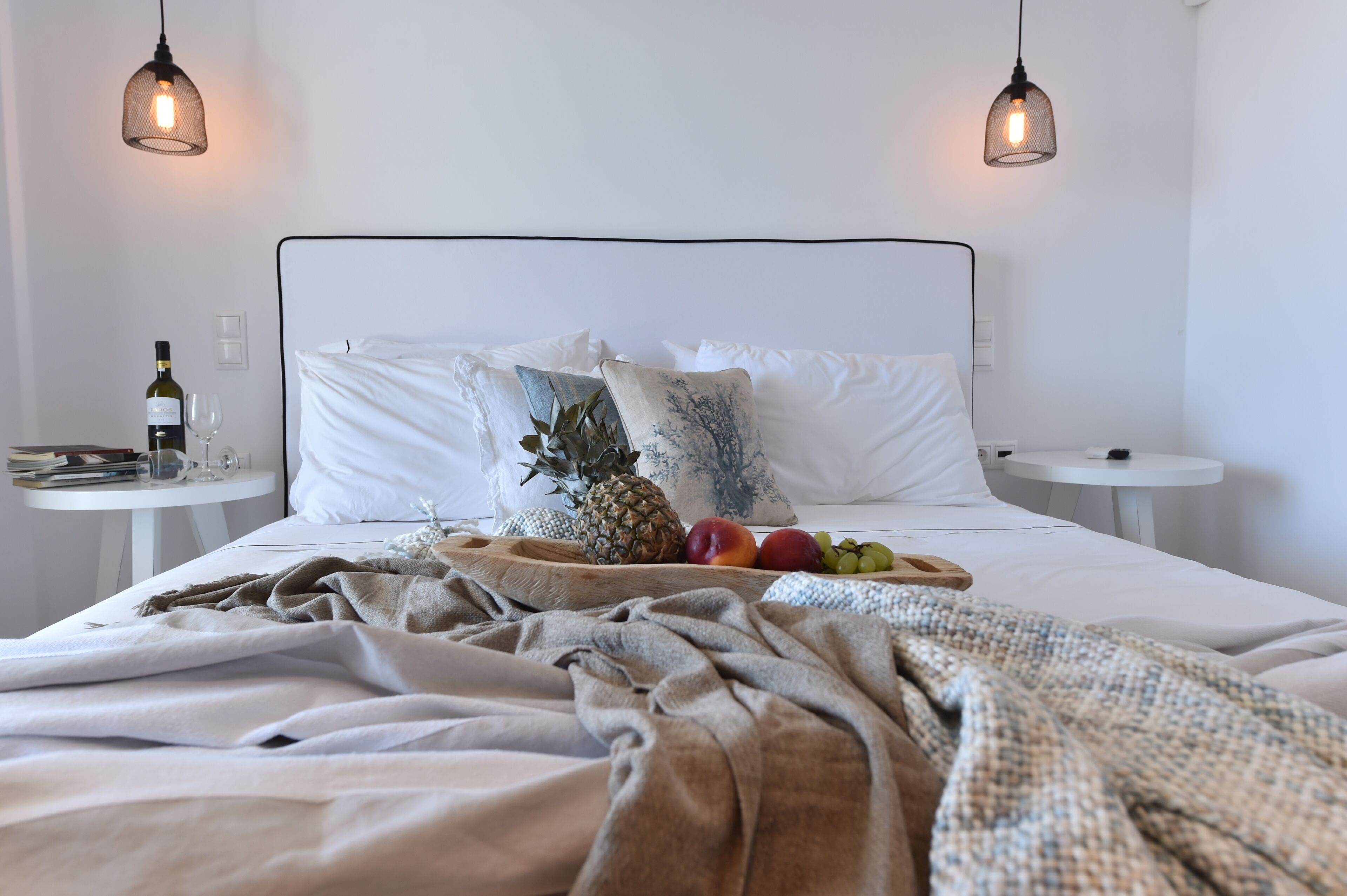 Una cama bien hecha con ropa blanca, un frutero con piña, uvas y melocotones, y dos mesitas de noche con una botella y copas en una.