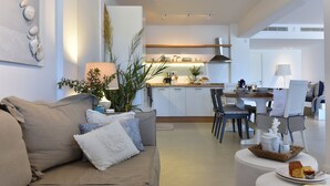 Uma sala de estar moderna com um sofá bege, uma área de jantar com mesa e cadeiras, e uma cozinha ao fundo.