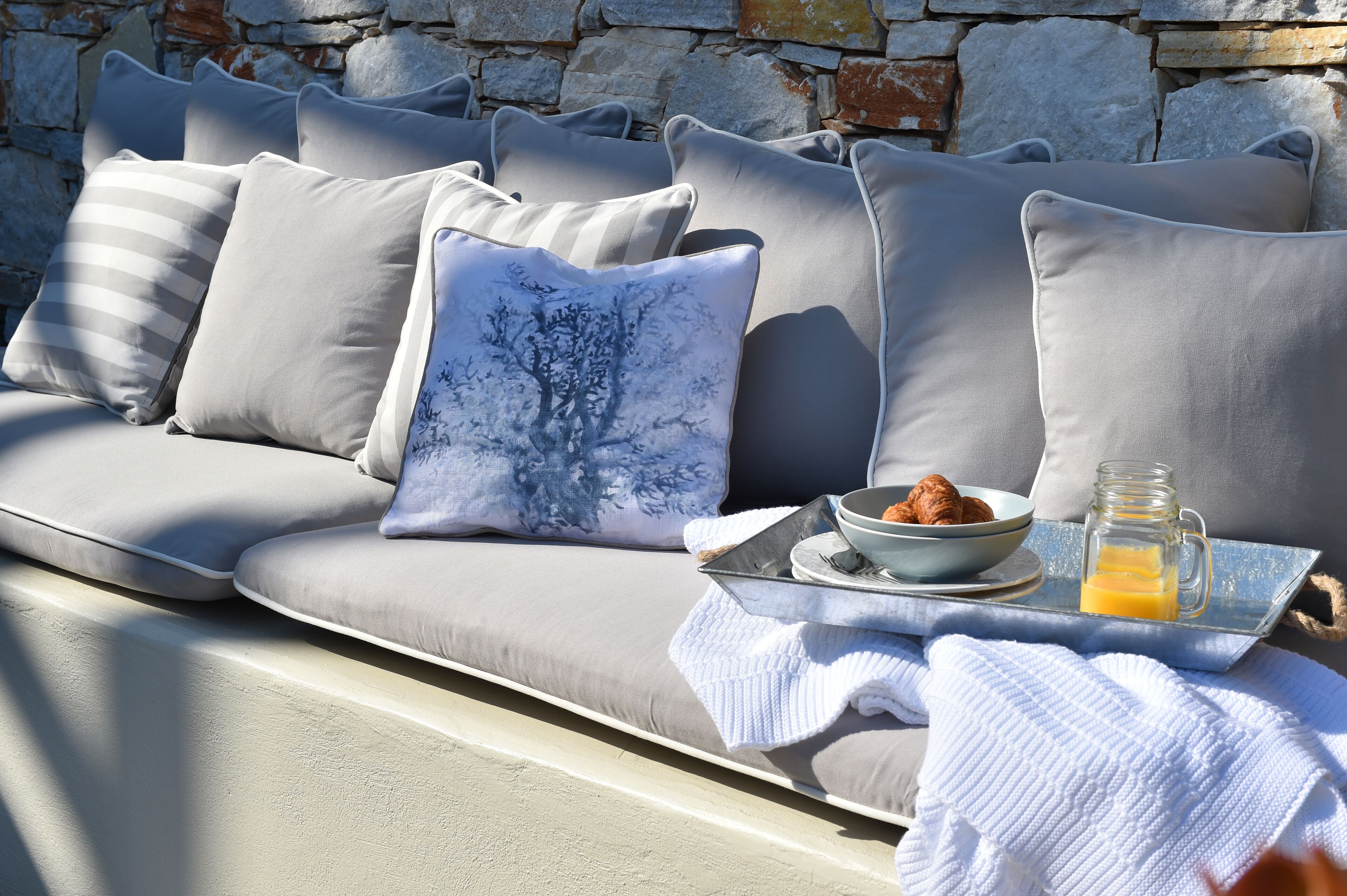 A modern outdoor seating area with a stone wall backdrop, a tray with croissants and orange juice, and a bowl of fruit.