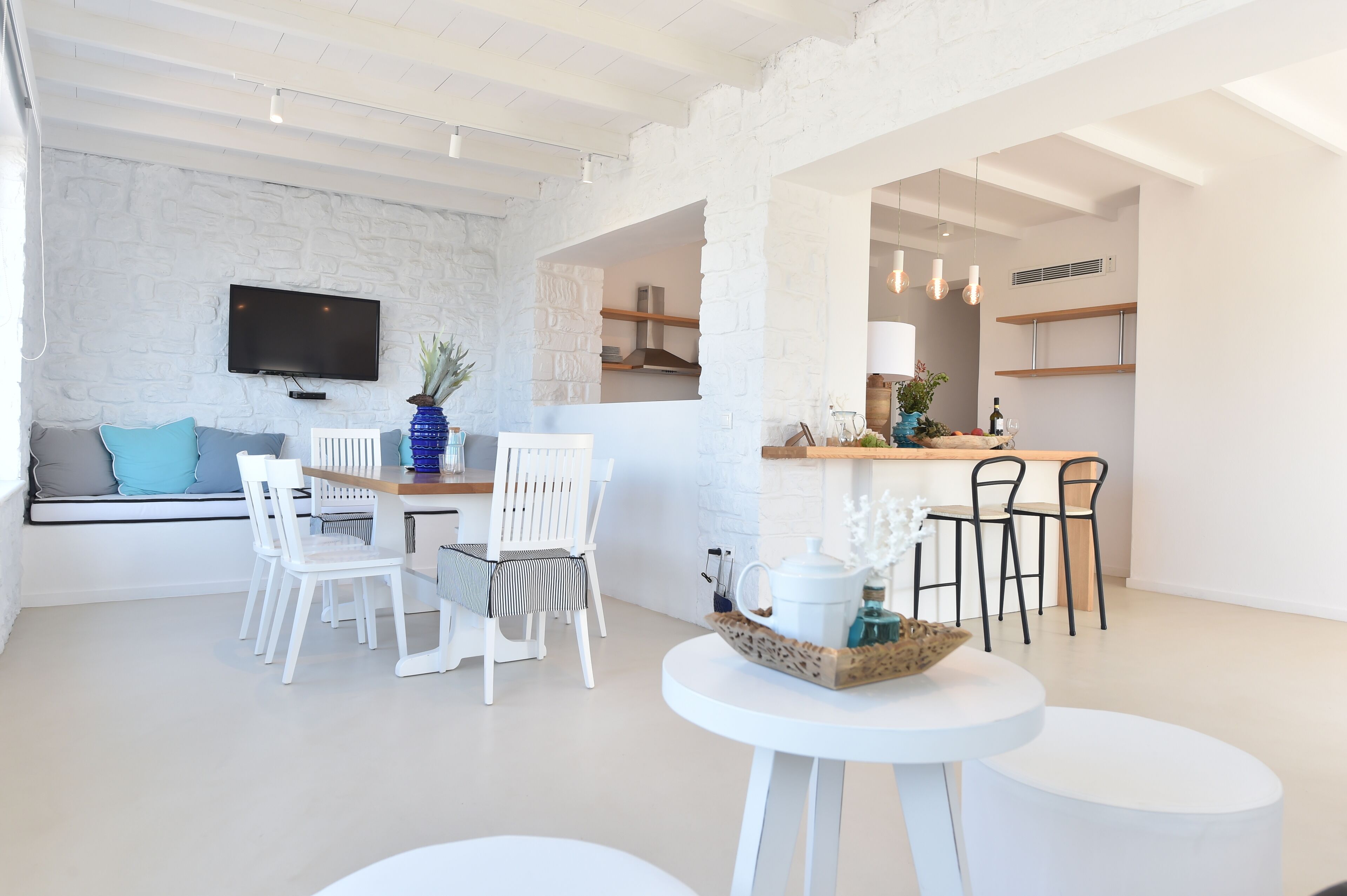 A modern interior with a white brick wall, a flat-screen TV, white dining chairs, a small round table, and a bar area with stools.