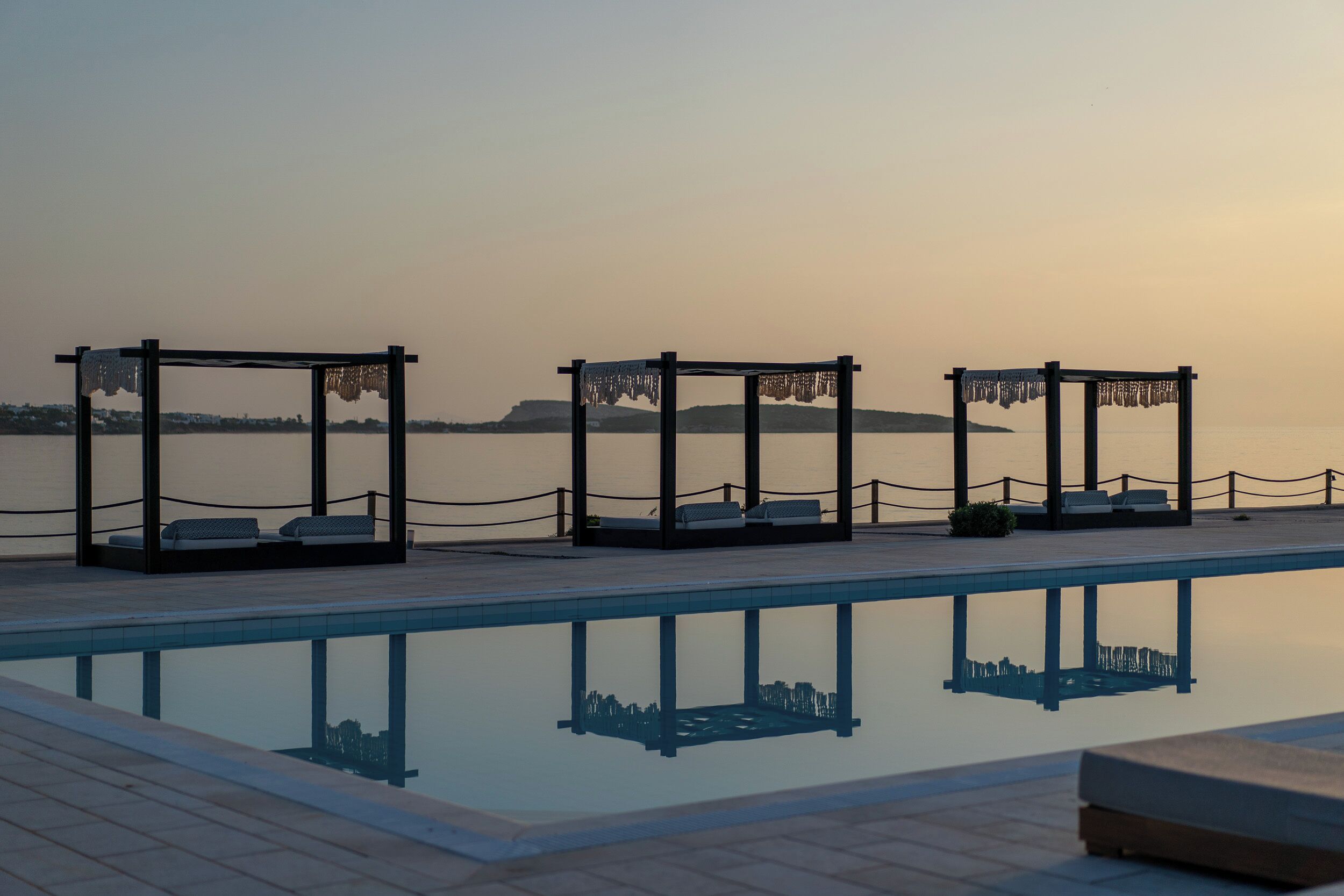 A poolside area with three wooden gazebos, each with a bench and a small table, overlooking a calm body of water.