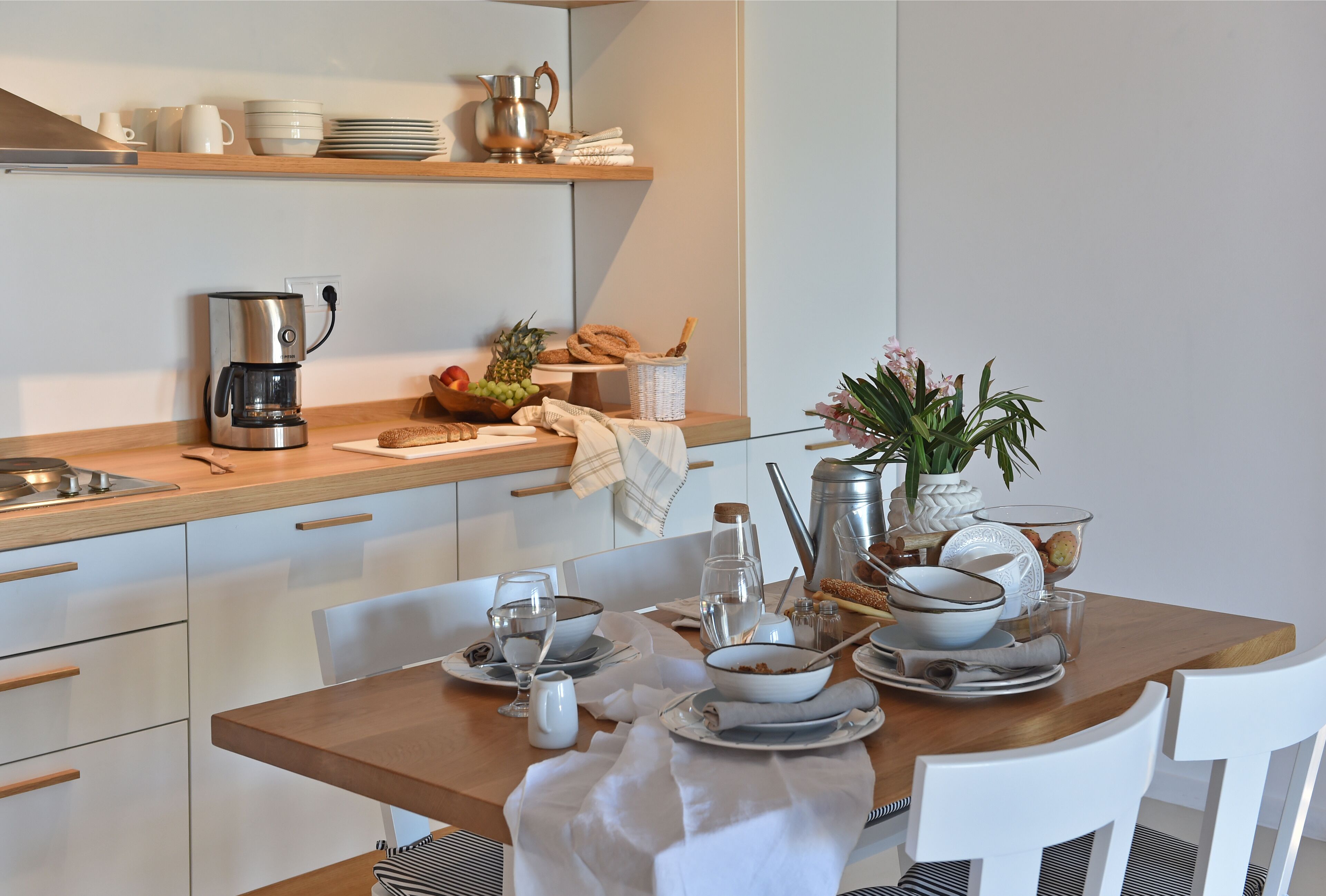 A modern kitchen with a wooden table set for breakfast, including plates, bowls, and a coffee maker.