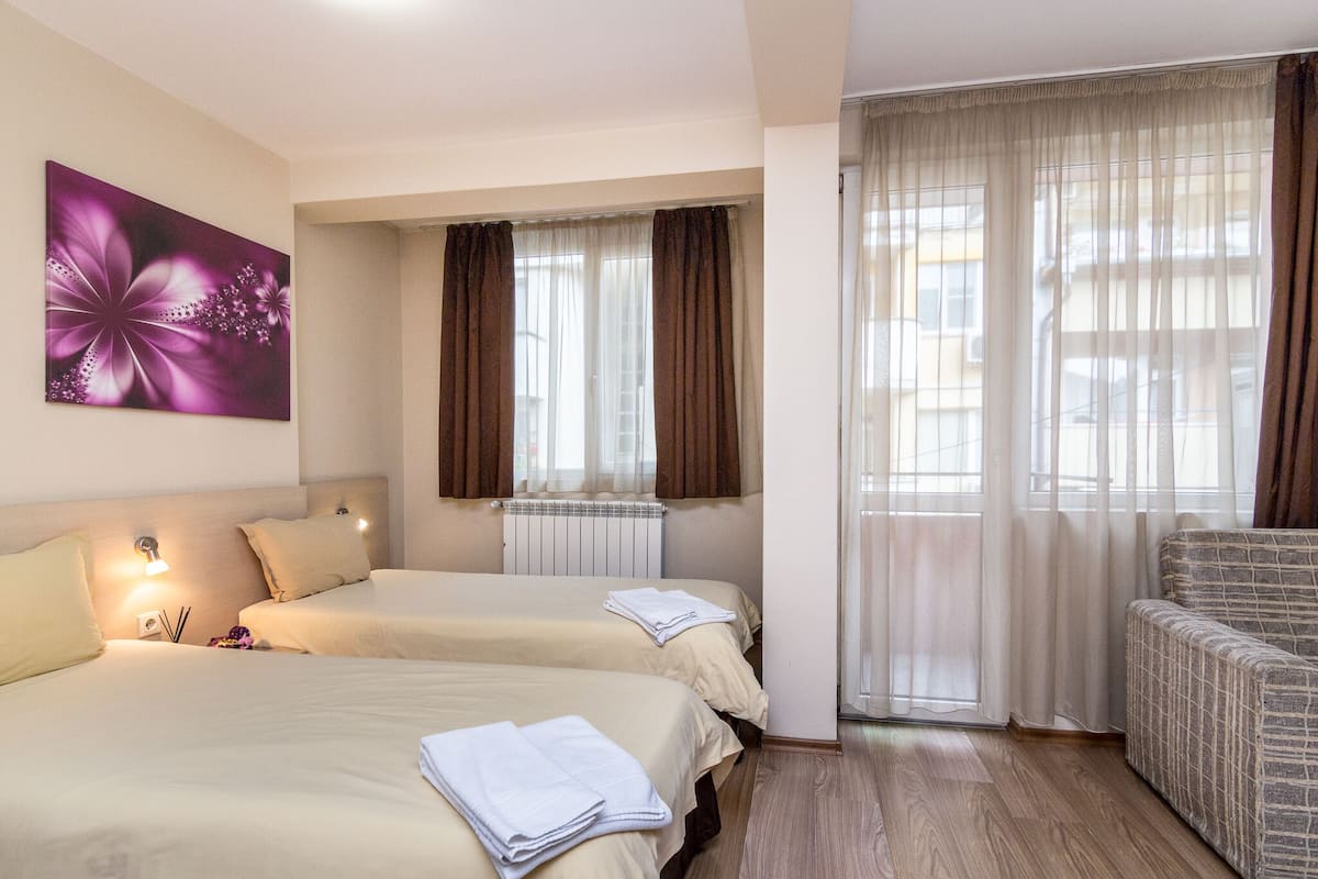 Habitación de hotel con dos camas, un sofá, un ventanal con cortinas, un radiador y una obra de arte colgada en la pared.