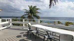 Una terraza con sillas y una mesa blancas, con vista al océano, palmeras y una barandilla blanca.