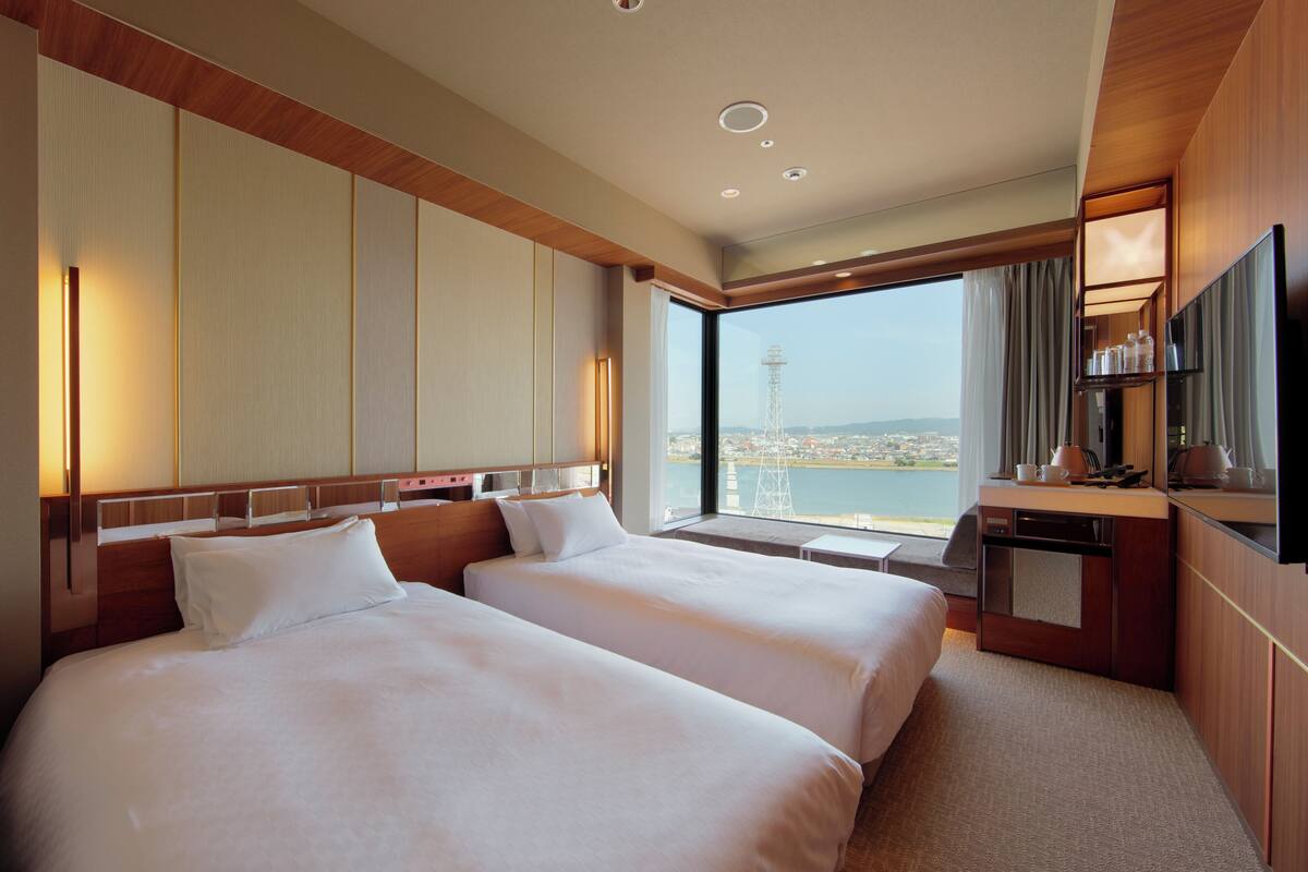 Habitación de hotel con dos camas, televisión, escritorio y vistas a un paisaje urbano.