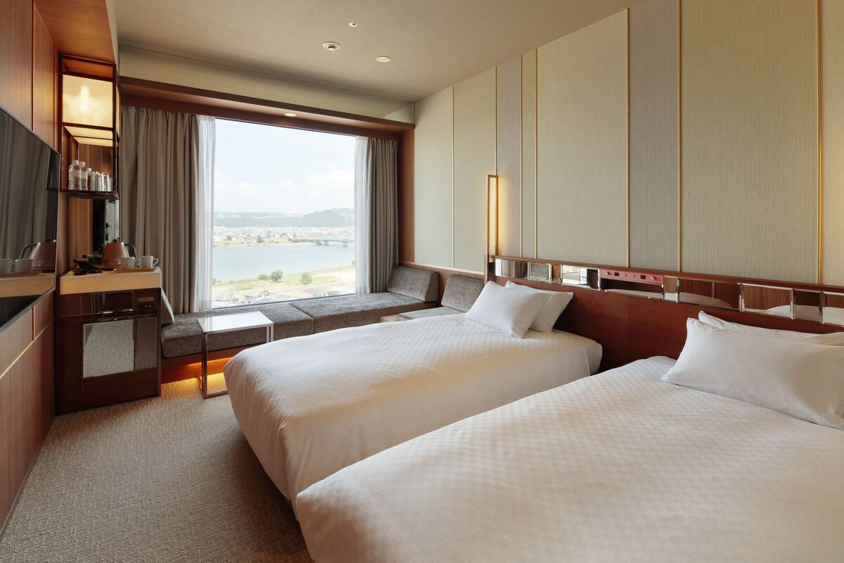Habitación de hotel con dos camas, un escritorio con espejo, una ventana con vistas al agua y un puente, y una pared grande con paneles verticales.