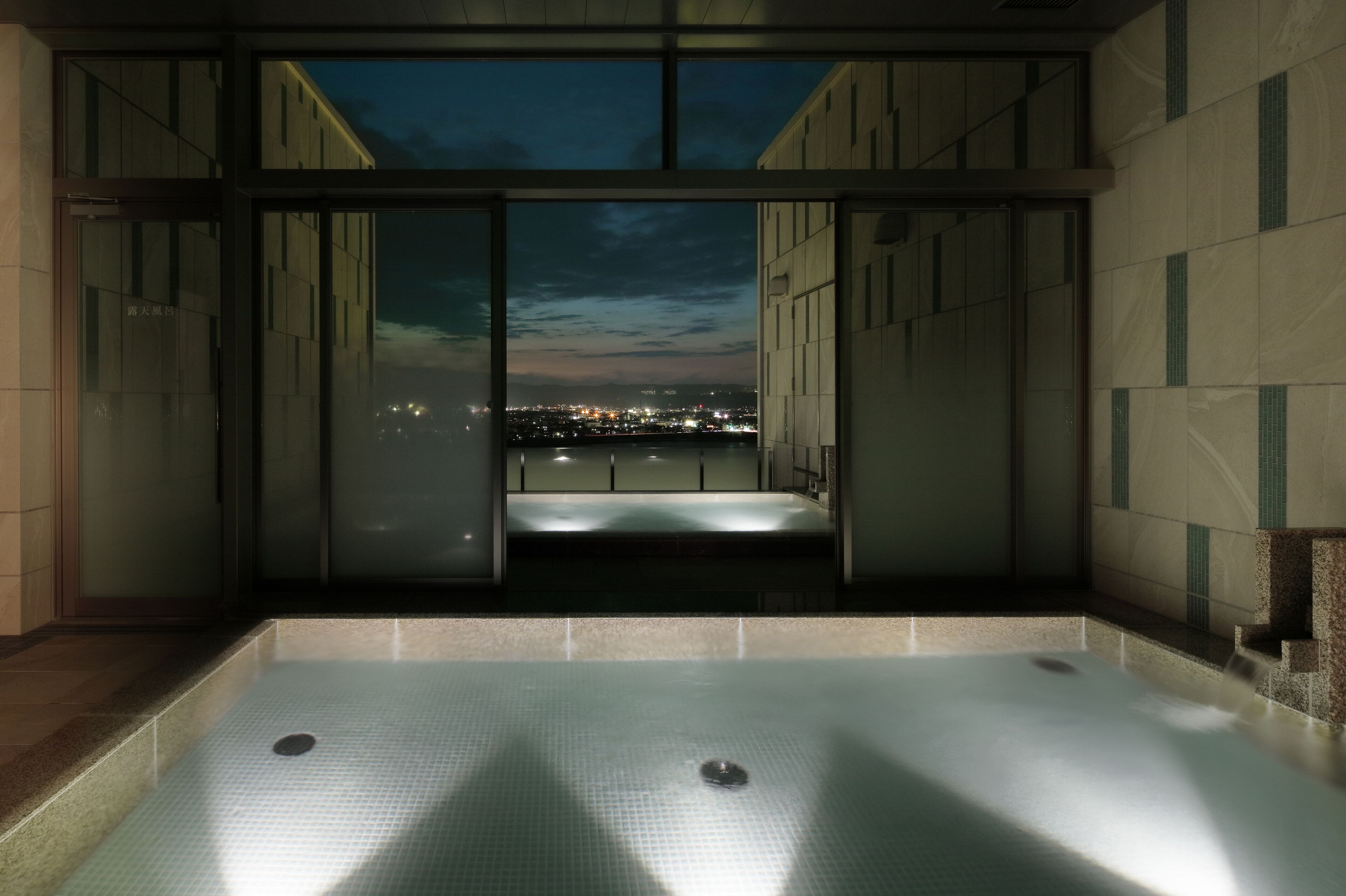 Área interna de piscina moderna com parede de vidro e vista para a cidade ao entardecer.