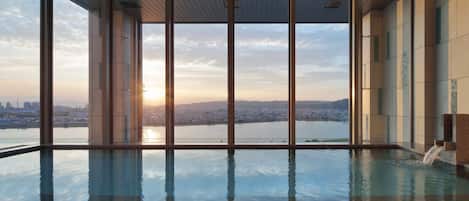 A modern indoor swimming pool with a city view, large windows, and a clear sky.