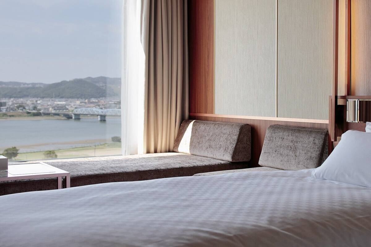 Habitación de hotel con una cama grande, zona de estar con vistas a un puente y al paisaje urbano, y un ventanal con cortinas.