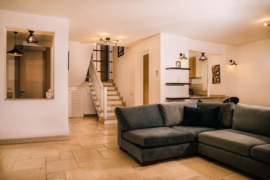 Un salon moderne avec un canapé d’angle gris, un escalier et un coin cuisine visible au fond.