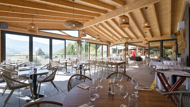 Un restaurante espacioso con vigas de madera y amplios ventanales que ofrecen vistas a la montaña. Las mesas están dispuestas con copas de vino y cubiertos.