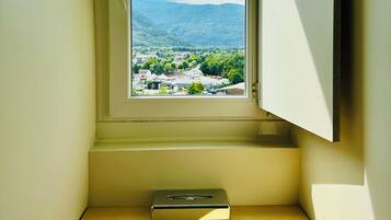 Una habitación pequeña con ventana que da a un valle, un escritorio con un libro y una estantería en la pared.