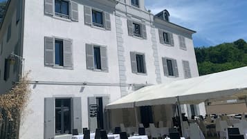 Un edificio blanco con múltiples ventanas y contraventanas, zona de estar exterior con mesas y sillas, y una gran sombrilla que da sombra.