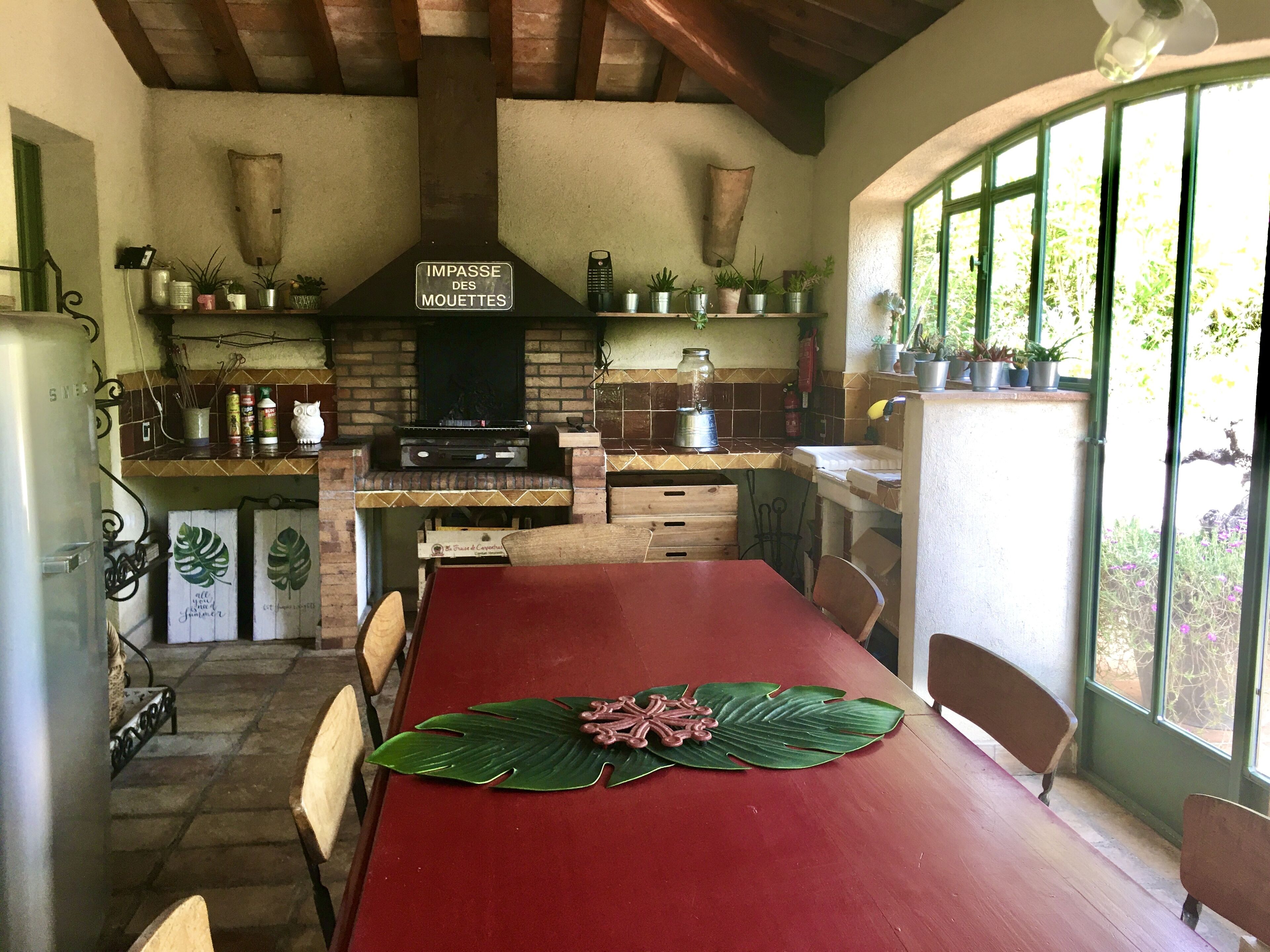 Een landelijke keuken met een bakstenen muur, houten kastjes en een ingebouwde grill. Er staat een eettafel met stoelen en een grote groene bladdecoratie.