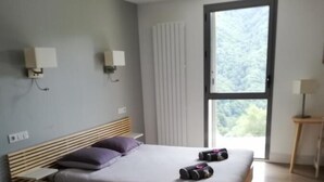 A modern bedroom with a wooden bed, a window with a view of greenery, and a small bedside table with a lamp. - Auberge des Isards (Aydius)