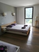 A modern bedroom with a wooden bed, a window with a view of greenery, and a small bedside table with a lamp.