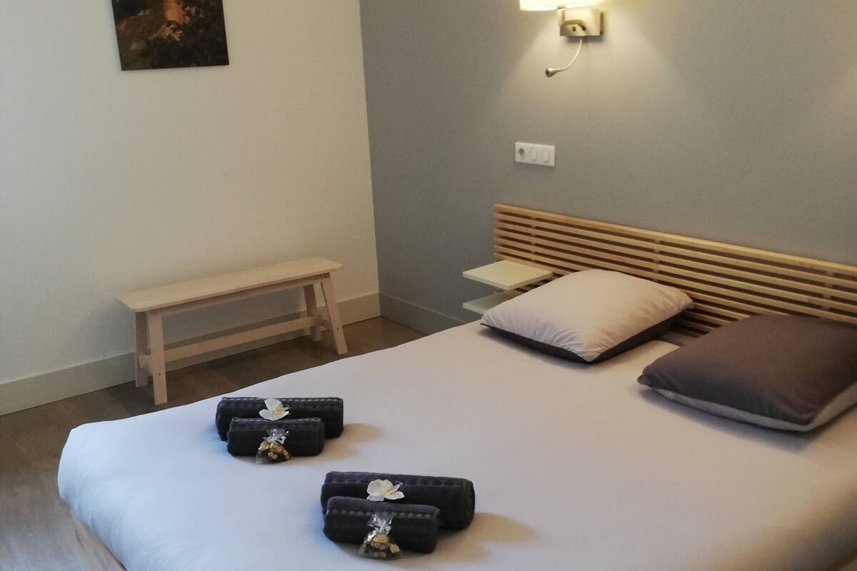 Una cama bien tendida con almohadas y dos toallas enrolladas, una mesita de noche, un banco y un aplique de pared.