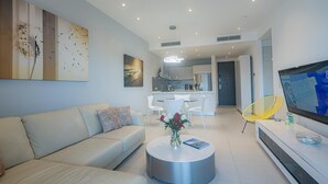 A modern living room with a sofa, coffee table, and a TV. There is a kitchen in the background with white cabinets and a dining area with white chairs. - K & E Lemon Drop Cove (Cupecoy)