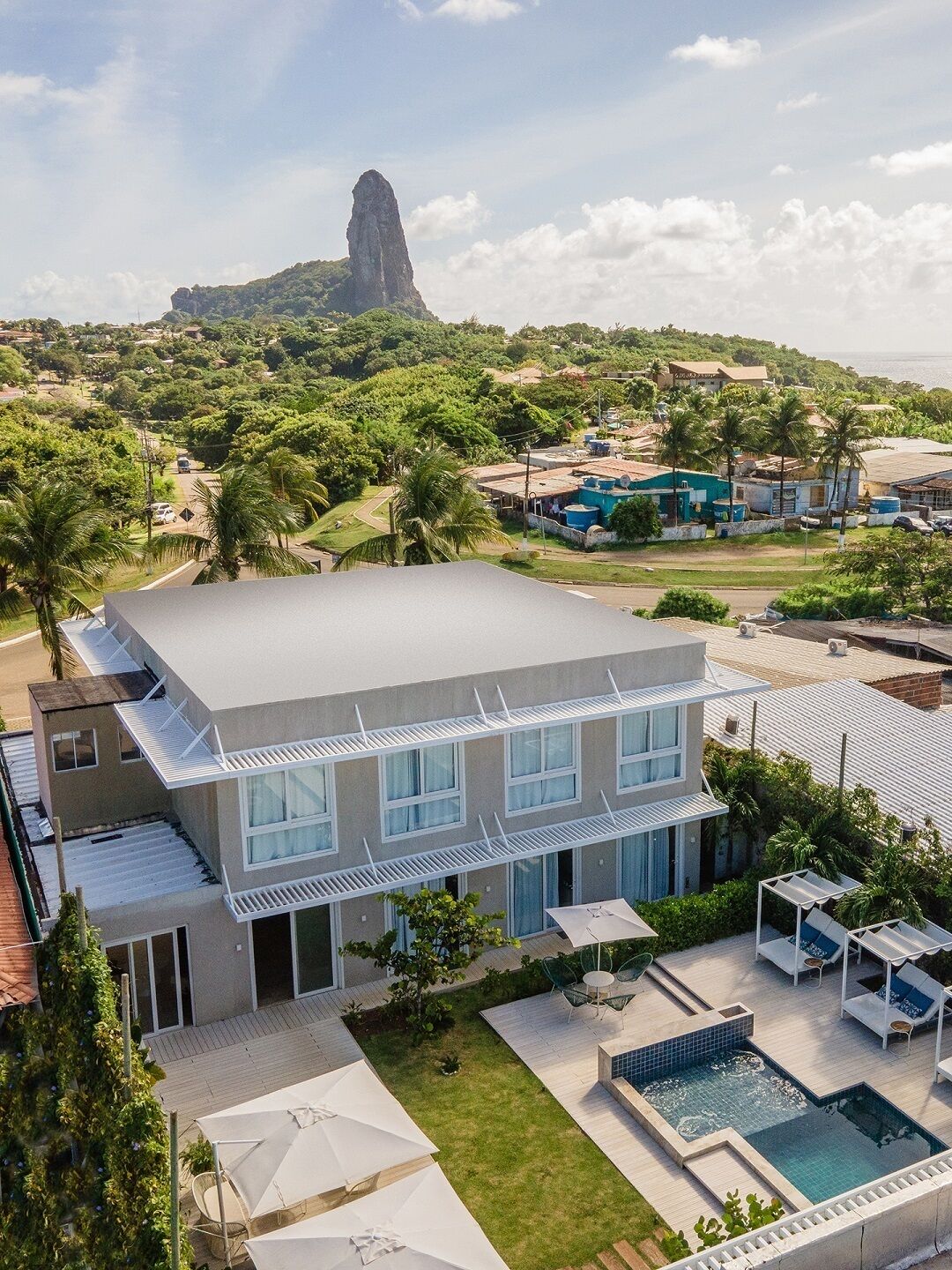 Um hotel moderno com piscina no terraço, rodeado por vegetação exuberante e uma montanha imponente ao fundo.