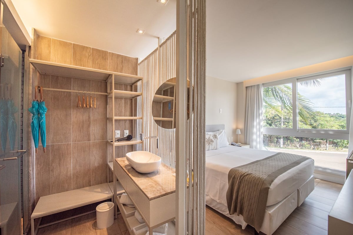 A modern bedroom with a large window, a bedside table, a bed, and a bathroom with a sink and shelves.